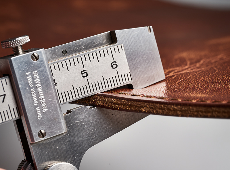 Macro close-up of a leather thickness gauge measuring a piece of vegetable-tanned wallet leather showing exactly 3 ounce or 1.2mm thickness on the dial