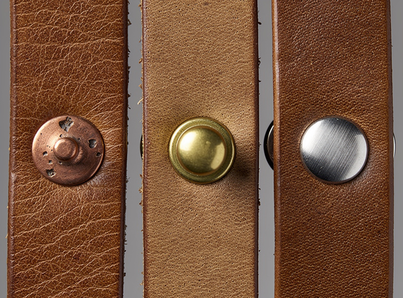 Macro close-up of three leather strap pieces fastened with different rivet types — left a copper burr rivet with hammered finish, middle a brass double-cap rivet with shiny dome, right a stainless steel single-cap rivet with brushed finish
