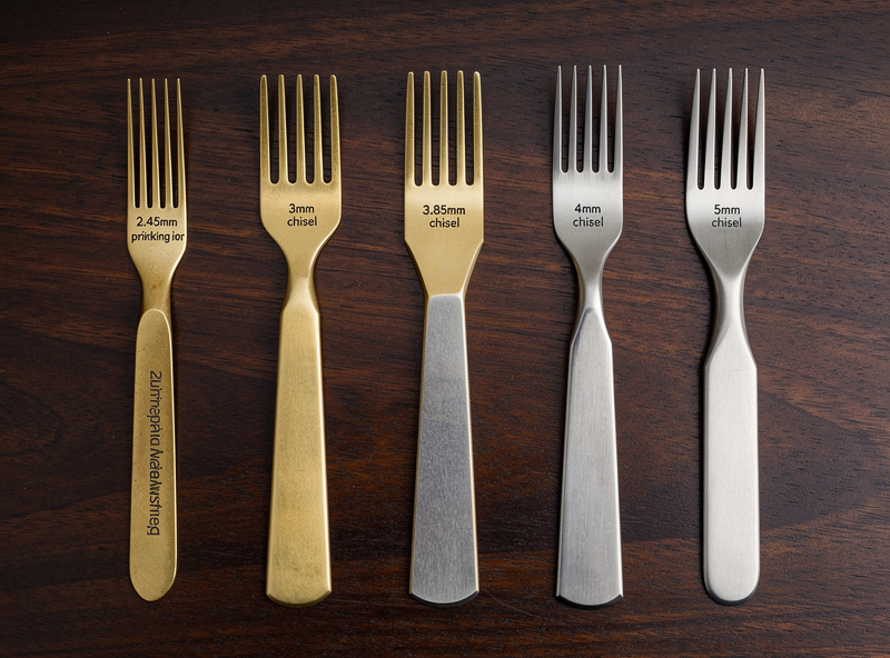 Five different stitching irons laid out side by side on dark wood showing different spacings: 2.45mm pricking iron, 3mm chisel, 3.85mm chisel, 4mm chisel, 5mm chisel