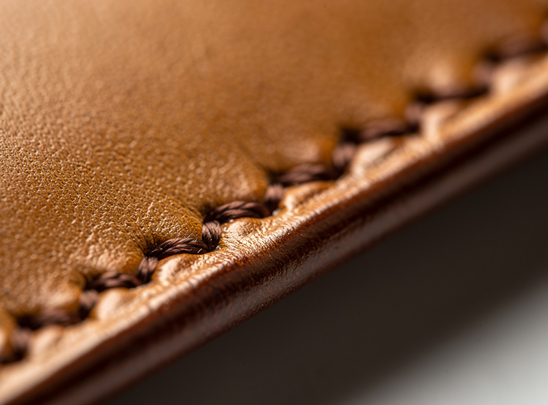Close-up of a finished hand-saddle-stitched leather wallet edge showing perfectly even diamond-shaped stitches with consistent angle and spacing in brown waxed thread on tan vegetable-tanned leather