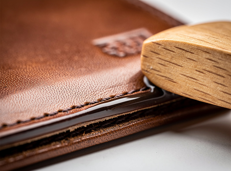 Close-up of a finished leather bifold wallet edge being burnished with a wooden slicker showing the leather transitioning from rough cut to glass-smooth polished surface with Tokonole compound visible