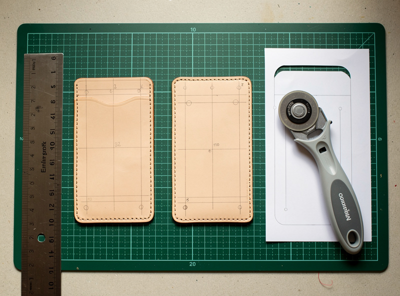 Three card holder leather panels cut from veg-tan stock with a steel ruler, rotary cutter, and paper template visible