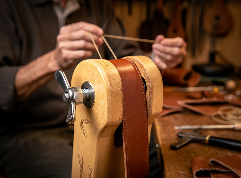Wooden stitching pony from the side with a leather belt clamped between jaws by a wing nut, person seated working with two needles