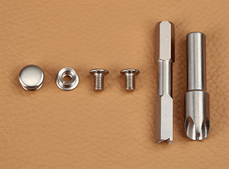Macro of a four-part snap set laid out on tan leather: cap, socket, post, stud, with steel anvil and concave setter punch beside them