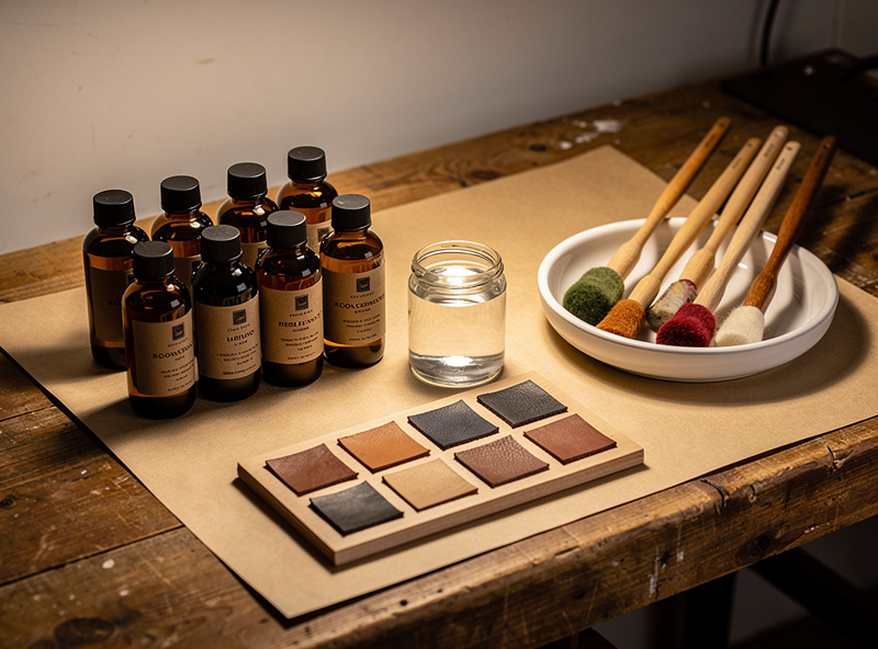 Workshop dye station with bottles of oil dye, alcohol dye, and water-based acrylic dye, wool daubers, and a leather sample swatch board