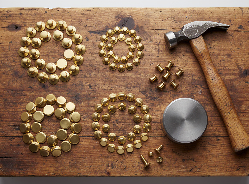 Assortment of solid brass leather rivets in different sizes lined up on a wooden workbench with a hammer and concave anvil die
