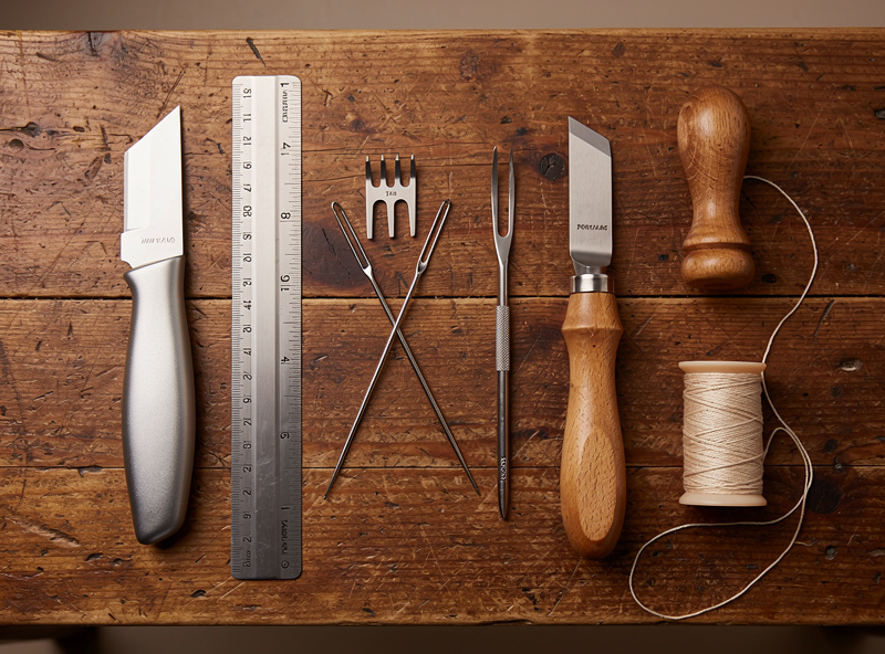 Organized leatherworking starter kit on a wooden workbench with knife, chisels, edge beveler, ruler, mallet, and thread spools