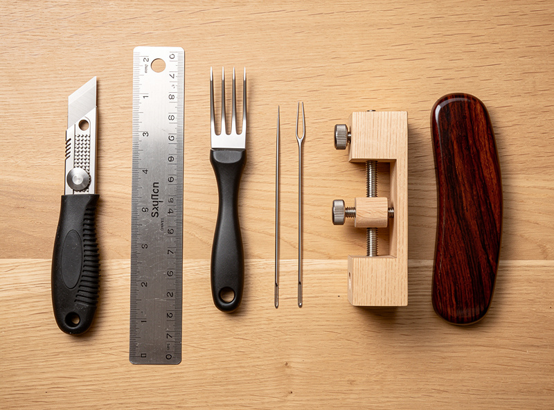 Six essential leatherworking tools laid out on a wooden workbench: utility knife with sharp blade, 12 inch steel ruler, four-prong stitching chisel, two harness needles, wooden stitching pony, and hardwood burnishing slicker