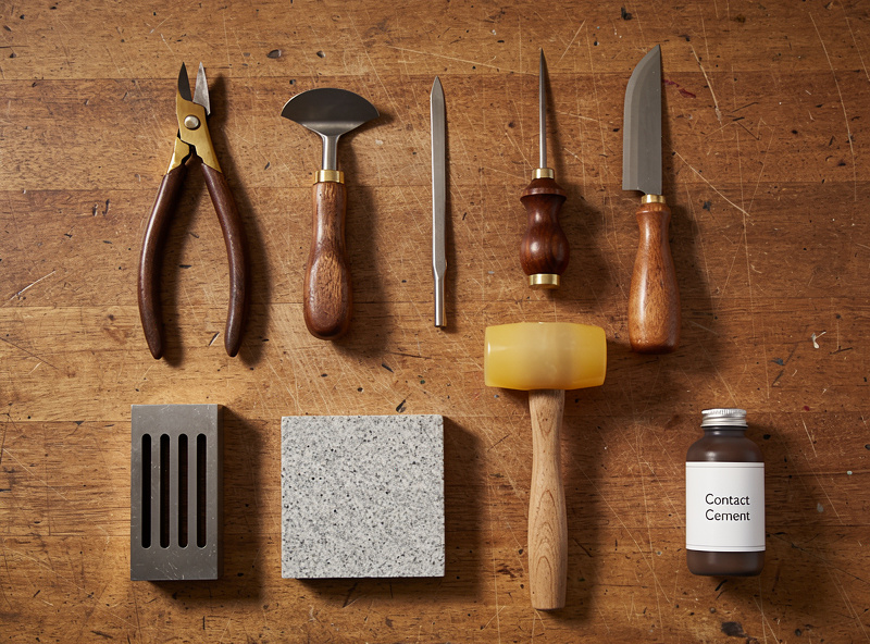 Mid-range leatherworking tool collection on a wooden workbench: wing divider, edge beveler, awl, skiving knife, slot punch, granite slab, polyurethane mallet, and a bottle of contact cement
