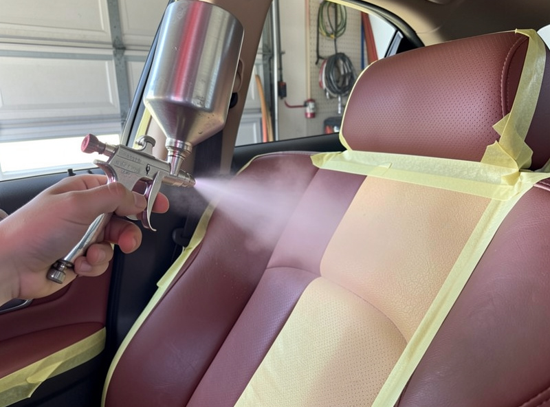 Spraying a color coat from an airbrush onto a prepared car leather seat with masking protecting the trim