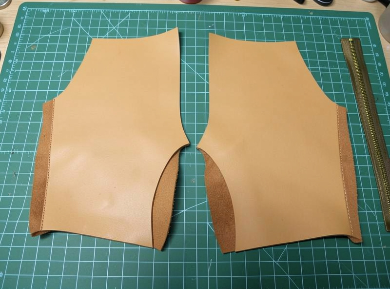Cut veg-tan body panel and two gusset pieces for a leather dopp kit laid out beside a brass zipper on a workshop bench