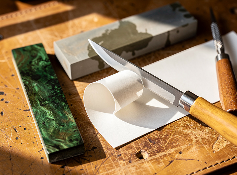 A head knife being tested by shaving a curl of paper after sharpening, with a charged strop, whetstone, and diamond awl arranged on a leather workbench in warm light