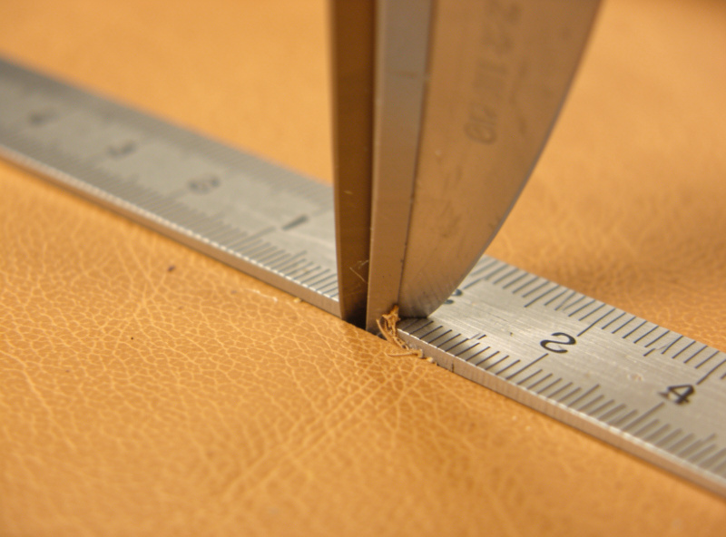 A vertical blade making a light scoring pass on thick leather against a steel ruler