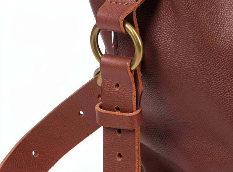 An adjustable leather bag strap with a roller buckle and punched holes attached to a bag through a D-ring