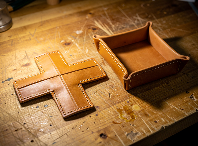 A flat cross-shaped piece of tan vegetable-tanned leather scored along fold lines beside a finished box-stitched valet tray on a workshop bench