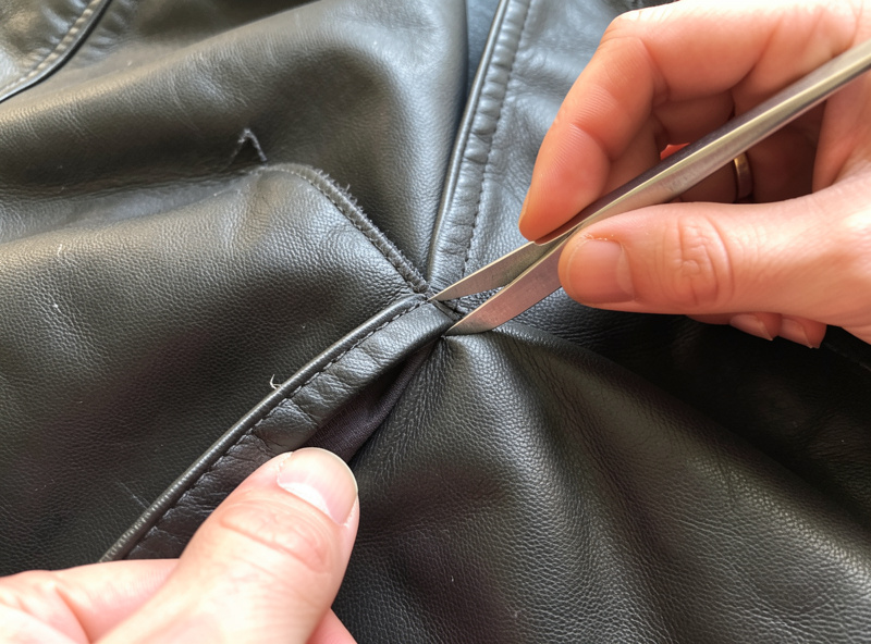 Hands using a seam ripper to carefully open the lining seam of a leather jacket, exposing the inside seam allowance