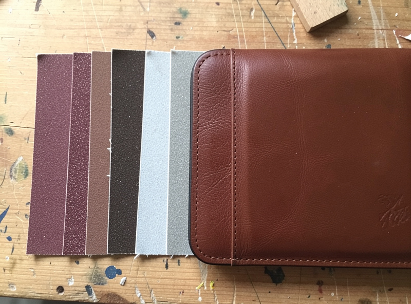 A row of sandpaper sheets in increasing grit beside a leather wallet edge being sanded
