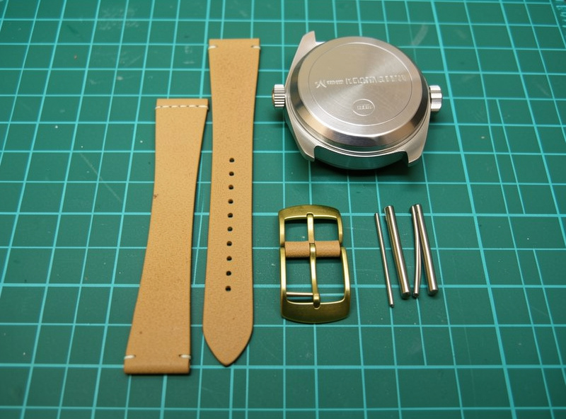Two cut pieces of 3-4 oz veg-tan watch strap leather next to a watch case, brass buckle, and spring bars on a workshop bench