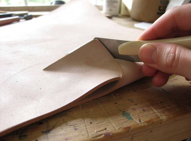 A folded turned edge on soft leather being pressed down with a bone folder, double-sided basting tape visible