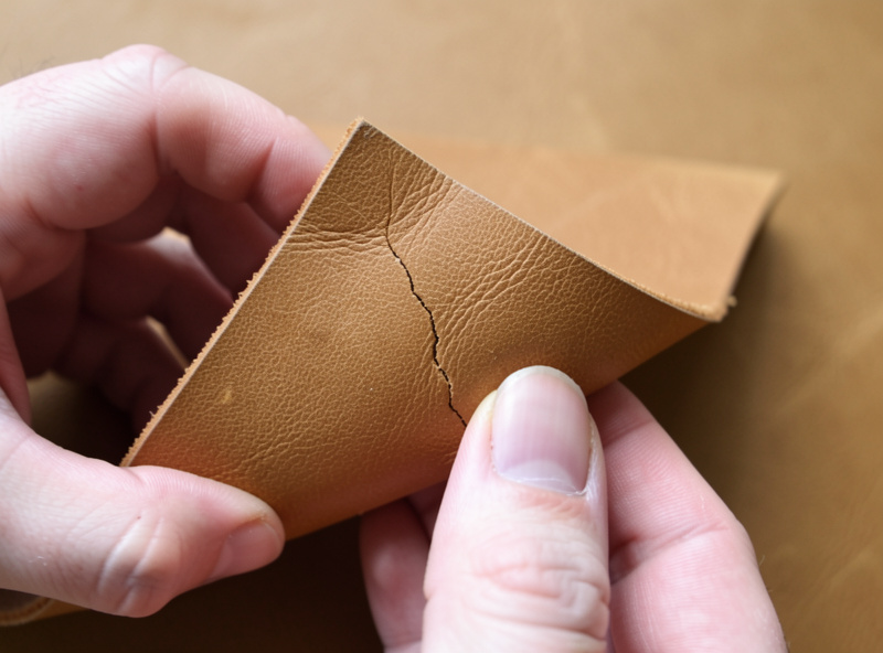 Hands flexing a folded corner of thick veg-tan leather showing a faint crack line on the grain