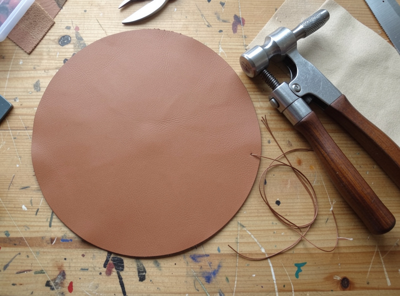 A flat circular leather blank beside a leather punch and waxed thread laid out on a workbench